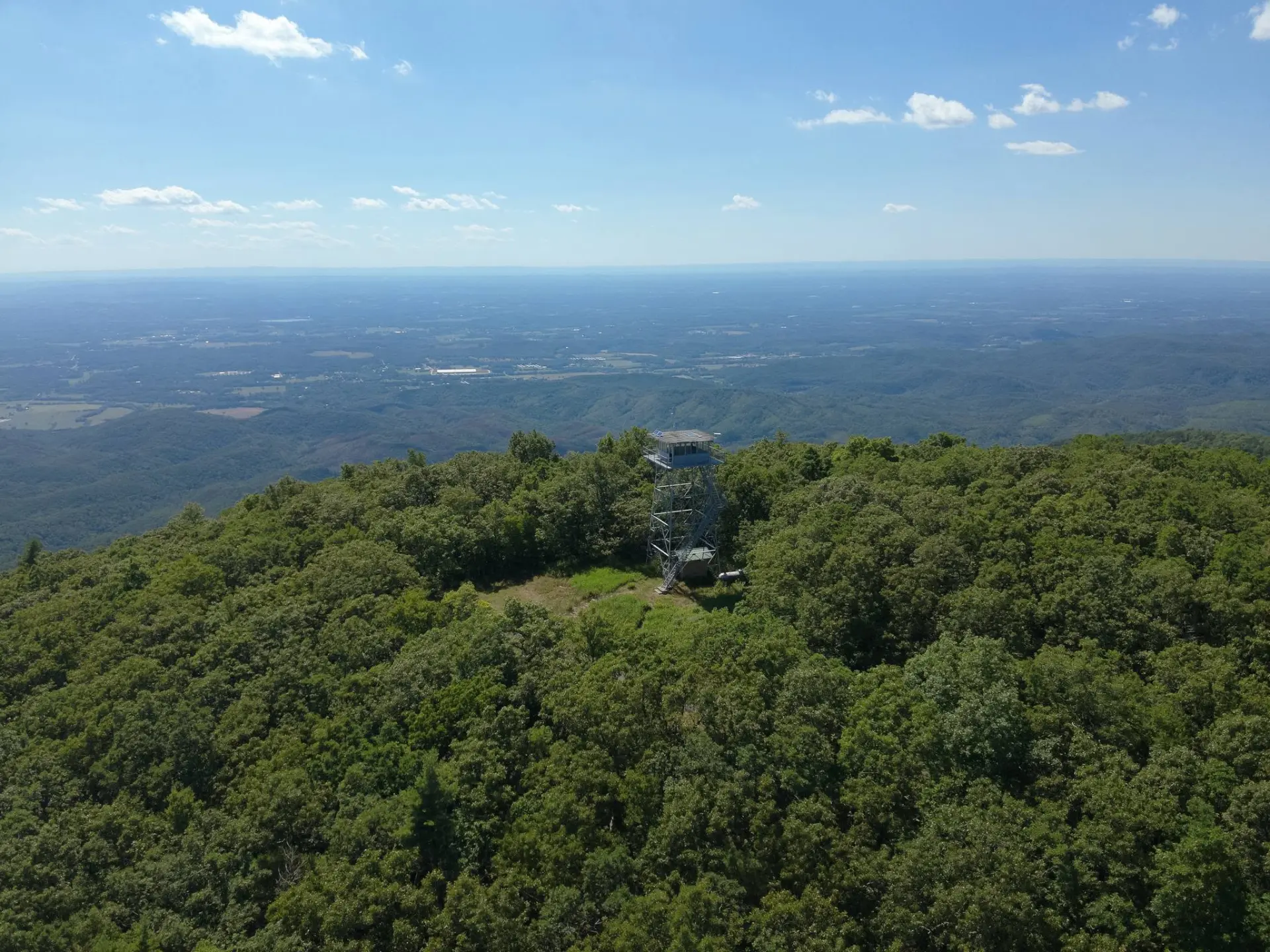 Aerial view of the Grassy Mountain fire tower rising above a dense green forest canopy, with ridgelines stretching to the horizon under a blue sky