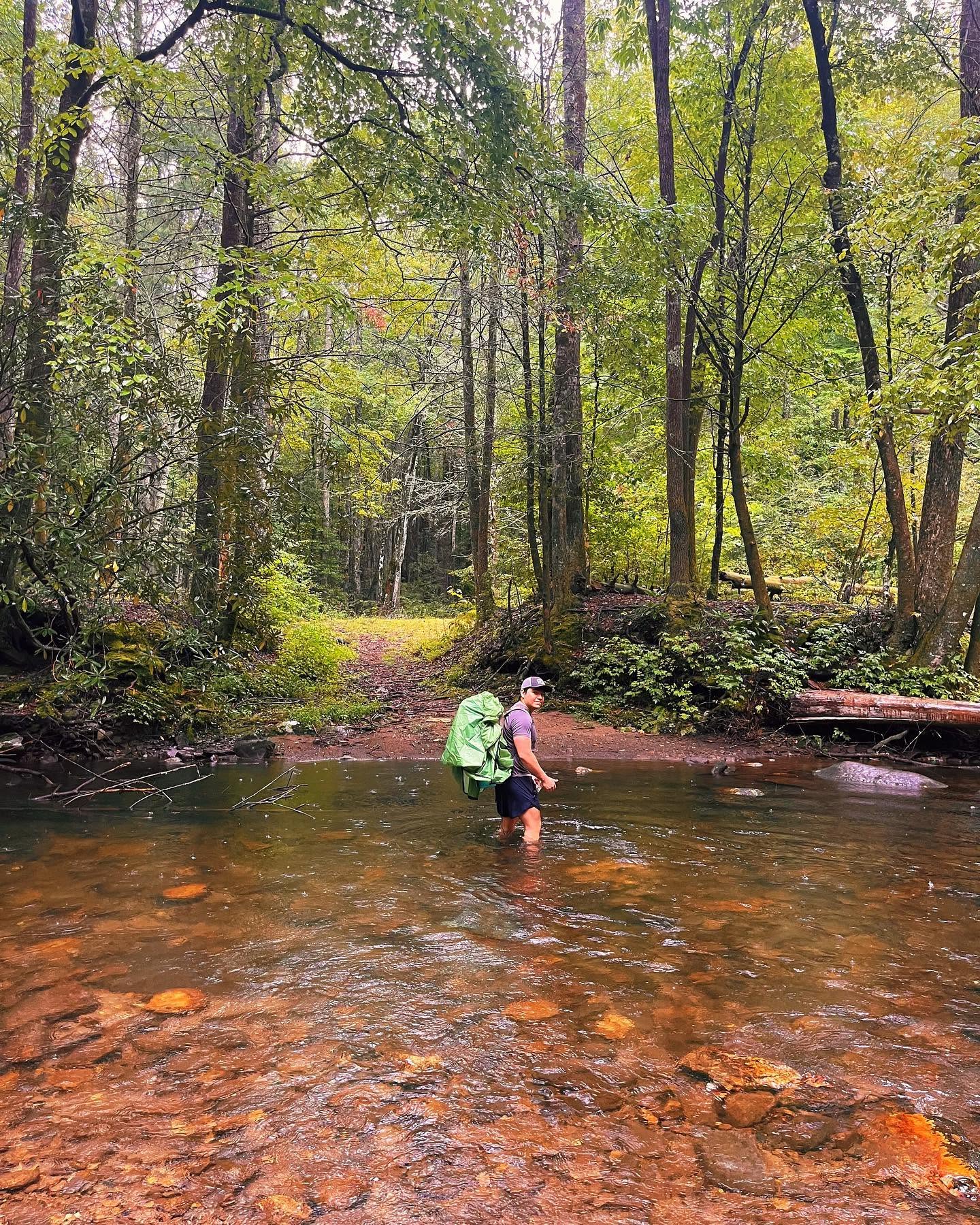 Backpacker with a green pack fording the shallow Conasauga River at Bray Field, surrounded by dense summer forest