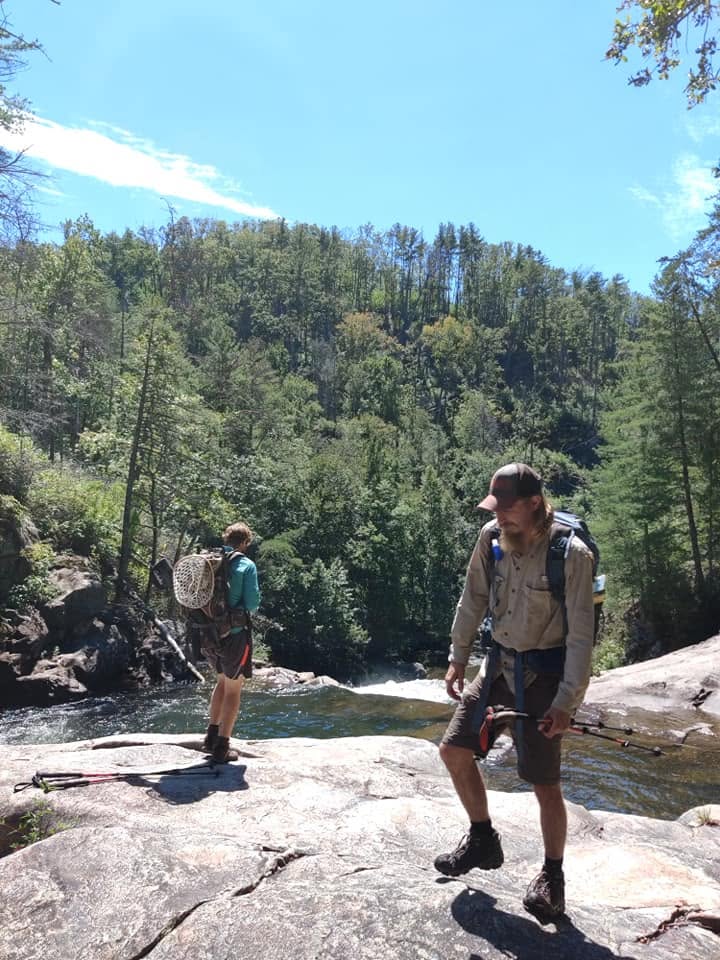 Two backpackers with trekking poles and a landing net standing on open bedrock above the Jacks River gorge on a clear day